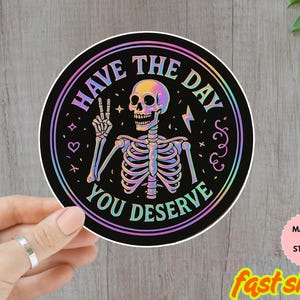May include: A round sticker with a black background and the text "HAVE THE DAY YOU DESERVE" in a rainbow gradient. A skeleton is in the center, giving a peace sign. The sticker is held by a hand.