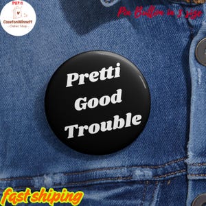 May include: A black pin-back button with the words "Pretti Good Trouble" in white, bold, sans-serif font. The button is set against a blue denim background. The button is round and appears to be about 2 inches in diameter.