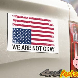 Può includere: Un magnete o adesivo rettangolare con un design invecchiato della bandiera americana. La bandiera ha strisce rosse e bianche e un campo blu di stelle. Il testo "WE ARE NOT OKAY" è stampato in nero sotto.