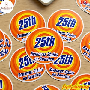 May include: Circular stickers with an orange and yellow concentric design. The stickers feature the number "25th" in blue, with the text "Removes Stains on America". The stickers are arranged on a wooden surface.