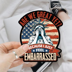 May include: A black and white sticker with a cartoon image of the Statue of Liberty with her hands over her face. The sticker says "Are we great yet? Because I just feel embarrassed." The background is a red, white, and blue American flag.