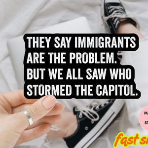 以下が含まれることがあります： 「THEY SAY IMMIGRANTS ARE THE PROBLEM. BUT WE ALL SAW WHO STORMED THE CAPITOL」というテキストが入った白黒のマグネットまたはステッカー。画像には指輪と黒いスニーカーも含まれています。