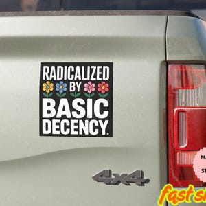 May include: A black and white magnet or sticker with the text "RADICALIZED BY BASIC DECENCY" and colorful flower graphics. The magnet is displayed on the back of a vehicle.