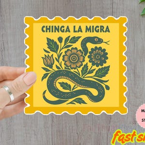 以下が含まれることがあります： 「CHINGA LA MIGRA」のテキストが入った、ヘビと花のモチーフが描かれた黄色の装飾ステッカー。ステッカーは手で持たれており、「WATER POOF」と表示されています。