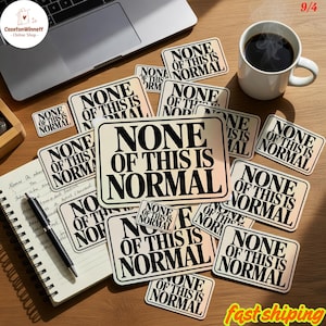 May include: A selection of rectangular stickers, each bearing the text "NONE OF THIS IS NORMAL" in black on a cream background. The stickers are arranged on a wooden surface, with a notebook, pen, and a coffee cup visible.