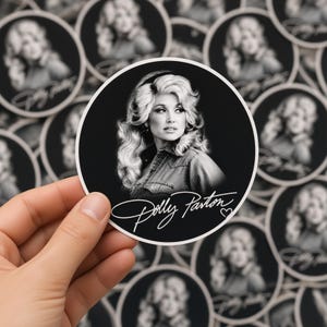 May include: A circular sticker featuring a black and white portrait of Dolly Parton with voluminous blonde hair, wearing a collared shirt. The name Dolly Parton is written in a white cursive script at the bottom with a small heart icon.