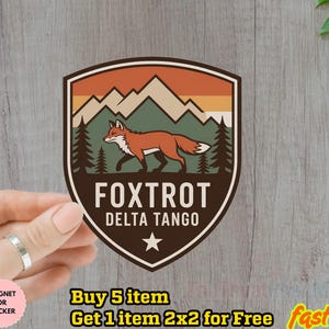 May include: A shield-shaped sticker featuring a fox walking across a mountain landscape. The sticker has a brown border with the words "FOXTROT DELTA TANGO" and a star at the bottom. The background includes mountains, trees, and a sunset.