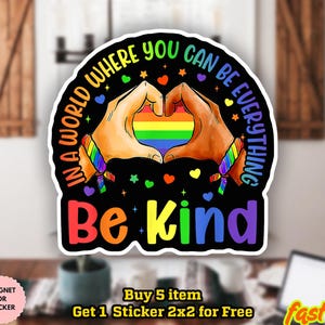 Puede incluir: Adhesivo negro con el texto "In a world where you can be everything, be kind". El diseño presenta manos formando un corazón alrededor de un arcoíris, con acentos coloridos. El texto es arcoíris y blanco.