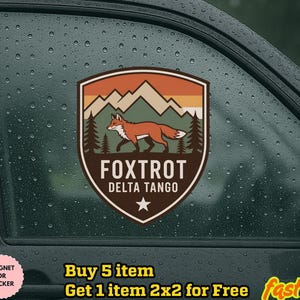 May include: A shield-shaped sticker featuring a fox walking in front of a mountain range and pine trees. The sticker has the text "FOXTROT DELTA TANGO" and a star at the bottom. The sticker is brown, orange, and green.