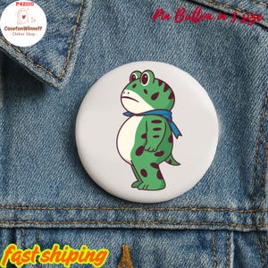 May include: A round pin button featuring a cartoon frog with green and brown spots, wearing a blue scarf. The button is set against a denim background. Text on the button reads "Pin Button in 1.25 inch".