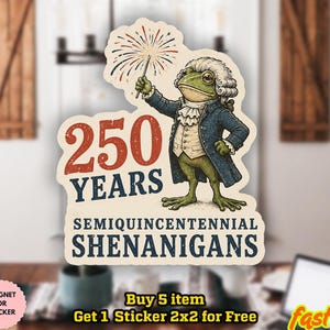 Könnte beinhalten: Ein gestanzter Aufkleber mit einem grünen Frosch in kolonialer Kleidung aus dem 18. Jahrhundert, der ein Feuerwerk hält. Der Text lautet 250 YEARS SEMIQUINCENTENNIAL SHENANIGANS in roter und blauer Schrift auf cremefarbenem Grund.