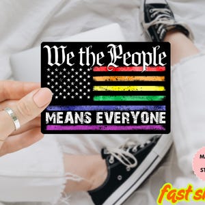 Può includere: Magnete o adesivo rettangolare nero con la scritta "We the People" in un font stilizzato. Il design incorpora la bandiera americana con strisce arcobaleno e le parole "MEANS EVERYONE".