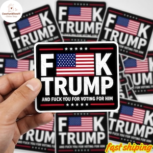 May include: A black sticker with the text "F**K TRUMP" in white, featuring a US flag design. The sticker also includes the phrase "AND F**K YOU FOR VOTING FOR HIM". Several similar stickers are visible in the background.
