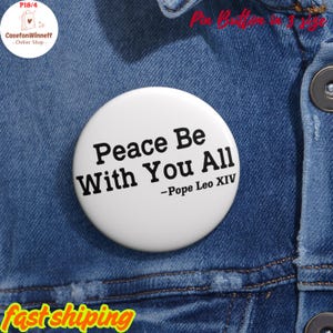 以下が含まれることがあります： 「Peace Be With You All - Pope Leo XIV」の黒い文字が書かれた白いボタン。青いデニムジャケットの上にあります。ボタンはCasetonWinnett Online Shopの商品です。