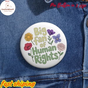 May include: A white pin button with the text "Big fan of Human Rights" in green and yellow, surrounded by colorful flower illustrations. The pin is set against a denim background.