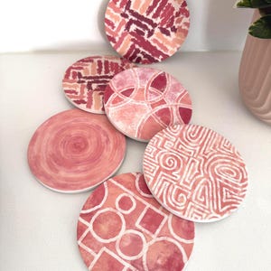 May include: Six circular coasters with abstract patterns in pink and red hues. Designs include brushstrokes, geometric shapes, and swirls. The coasters are displayed on a white surface, highlighting their decorative nature. Each coaster is approximately 10 cm in diameter.