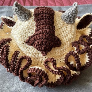 May include: A crocheted stuffed animal shaped like a mythical creature with brown and cream colored fur, two horns, and a curly mane.