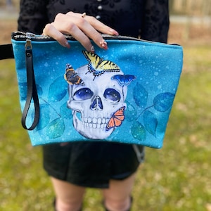 May include: A blue canvas zippered pouch with a skull design and colorful butterflies. The skull has a white background and the butterflies are yellow, orange, and blue.
