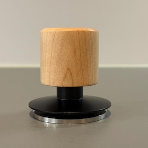 Könnte beinhalten: Ein Holz-Tamper mit einem zylindrischen hellbraunen Griff und einem schwarzen Sockel. Der Sockel hat ein kreisförmiges Design mit silbernem Rand. Der Tamper ist für Kaffee oder Espresso konzipiert.
