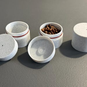 Könnte beinhalten: Ein Satz kleiner, hellgrauer Behälter mit Deckeln. Ein Behälter ist geöffnet und mit Kaffeebohnen gefüllt. Jeder Behälter hat ein rotes Band um die Mitte. Die Deckel haben ein kleines Loch in der Mitte.