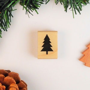 May include: A wooden stamp with a black silhouette of a Christmas tree. The stamp is on a white background with green pine branches and a brown wooden ornament.