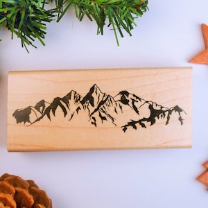 May include: A rectangular wooden stamp featuring a black mountain range design. The stamp is made of light-colored wood and is ideal for crafting or adding a nature-inspired touch to projects. The mountain range is a detailed silhouette.