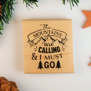 May include: A wooden stamp with the text "The Mountains are Calling & I Must Go" in black lettering. The stamp features a mountain range with a banner above it and pine trees below.