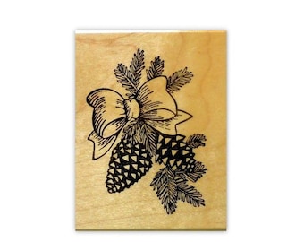 May include: A rectangular wooden rubber stamp featuring a black ink illustration of two pinecones and evergreen sprigs tied together with a decorative bow. The natural light-colored wood block has a smooth finish and a clear, detailed design.