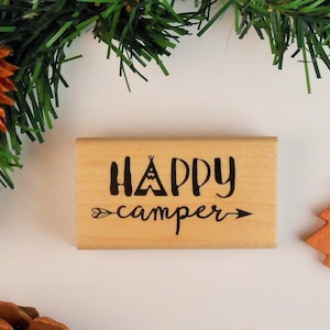 May include: Wooden stamp with the text "HAPPY camper" in black lettering. The text includes a small tent graphic and an arrow.