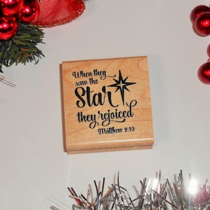 May include: A wooden stamp with the text "When they saw the star they rejoiced Matthew 2:10". The text is in black and the background is a light wood color. There is a star with a white center and black rays.