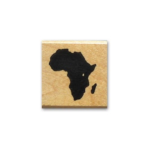 May include: A square wooden block featuring a black silhouette of the African continent. The wood has a light brown hue. The continent's shape is clearly defined, with its various geographical features.