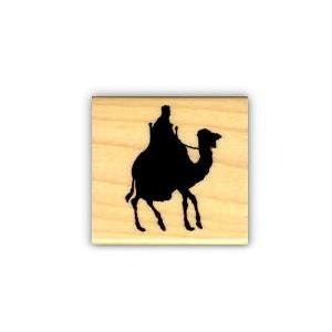 May include: Black silhouette of a person riding a camel. The image is a woodblock stamp.
