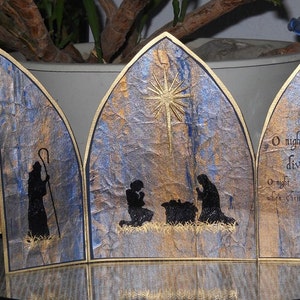 Op de afbeelding: Een driedelige kerstkaart met een gouden en blauwe textuur achtergrond. Het middelste paneel toont de kerststal met een ster erboven. Het linker paneel heeft de tekst "Peace on Earth" en het rechter paneel heeft de tekst "O night divine. O night when Christ was born."