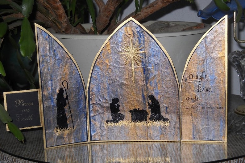 Op de afbeelding: Een driedelige kerstkaart met een gouden en blauwe textuur achtergrond. Het middelste paneel toont de kerststal met een ster erboven. Het linker paneel heeft de tekst "Peace on Earth" en het rechter paneel heeft de tekst "O night divine. O night when Christ was born."