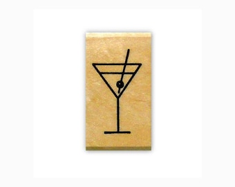 May include: A rectangular wooden block stamp featuring a black line art illustration of a martini glass with a single olive on a toothpick. The stamp has a light tan, natural wood grain finish and a smooth, flat surface for clear impressions.