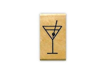 May include: A rectangular tan wooden stamp featuring a black line art illustration of a martini glass with a single olive on a toothpick inside the glass. The design is centred on the flat surface of the block.