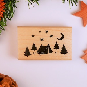 May include: A wooden camping-themed stamp featuring a black tent, trees, stars, and a crescent moon. The stamp is rectangular and made of light-colored wood. Decorative wooden star and tree shapes are nearby.