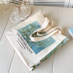 Peut inclure: Sac fourre-tout en toile écrue avec l'œuvre "Amandiers en fleurs 1890" de Vincent Van Gogh. Le sac est rectangulaire avec des anses blanches et un imprimé floral bleu et blanc. Le texte "Vincent Van Gogh" est imprimé.