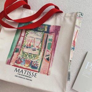 May include: Cream canvas tote bag with red handles. The bag displays a vibrant print of Matisse's "The Open Window" painting. Text on the bag reads "MATISSE <THE OPEN WINDOW> 1905 CANVAS OIL PAINTING". A small book is next to the bag.
