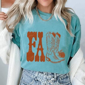 May include: Teal-colored t-shirt with the word "FALL" in orange, stylized text. The shirt features an orange illustration of cowboy boots and a pumpkin, evoking an autumn theme.