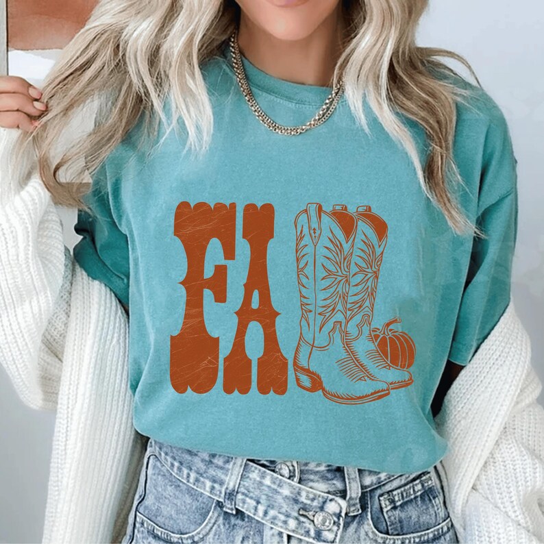 May include: Teal-colored t-shirt with the word "FALL" in orange, stylized text. The shirt features an orange illustration of cowboy boots and a pumpkin, evoking an autumn theme.