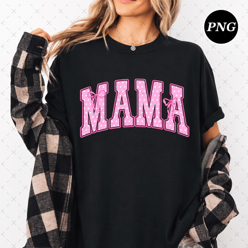 Pode incluir: Uma t-shirt preta com a palavra MAMA numa fonte universit&aacute;ria rosa com bolinhas brancas e um contorno rosa escuro. As letras M e A s&atilde;o decoradas com pequenos la&ccedil;os cor-de-rosa. A t-shirt &eacute; usada por baixo de uma camisa de xadrez preta e bege.