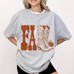 May include: A light gray sweatshirt with a brown graphic that says "FA" and features a pair of cowboy boots and a pumpkin. The boots have intricate floral designs.