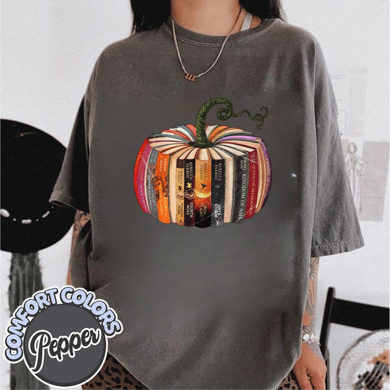 May include: Dark gray t-shirt featuring a colorful pumpkin design made of books. The pumpkin has a green stem and is surrounded by a variety of book spines in shades of red, orange, and yellow. The shirt has the words "COMFORT COLORS Pepper" printed on it.