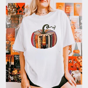 May include: White t-shirt featuring a pumpkin design made of books. The pumpkin has a green stem and is composed of various book spines in shades of orange, red, and brown. The shirt is a casual style, suitable for fall or Halloween.