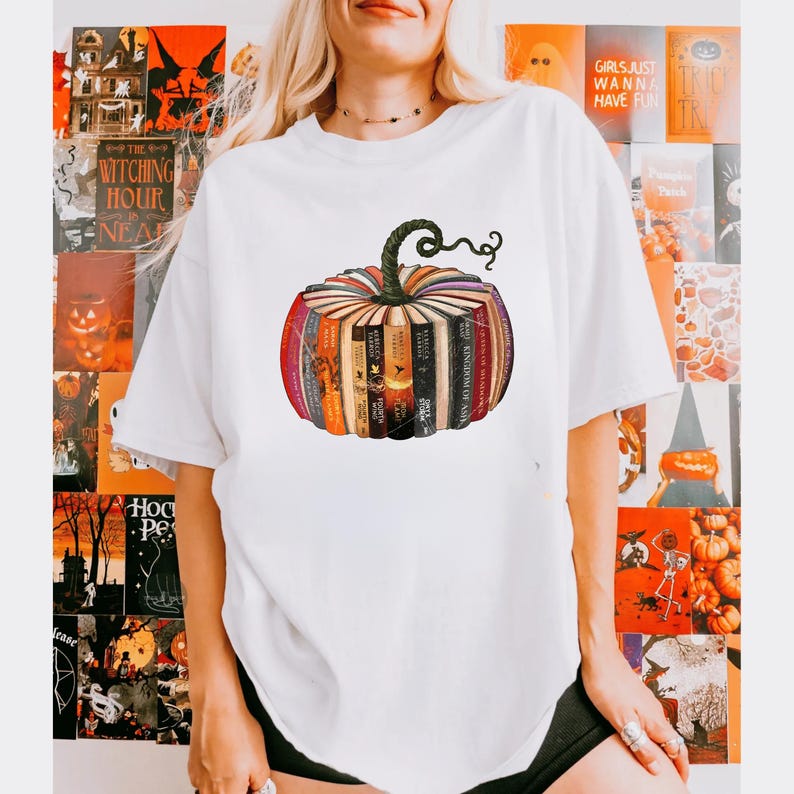 May include: White t-shirt featuring a pumpkin design made of books. The pumpkin has a green stem and is composed of various book spines in shades of orange, red, and brown. The shirt is a casual style, suitable for fall or Halloween.