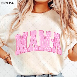 Pode incluir: Uma t-shirt de manga curta cor de creme com a palavra MAMA em letras grandes de estilo bloco cor-de-rosa com um padr&atilde;o de bolinhas brancas. A primeira e a &uacute;ltima letra est&atilde;o decoradas com pequenos la&ccedil;os cor-de-rosa desenhados &agrave; m&atilde;o.