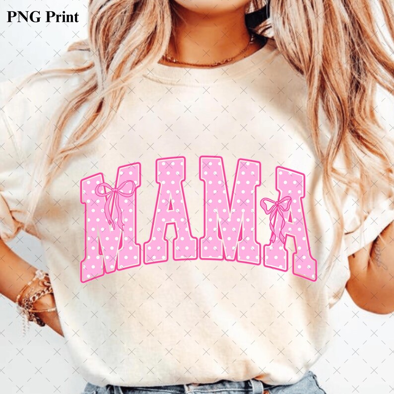 Pode incluir: Uma t-shirt de manga curta cor de creme com a palavra MAMA em letras grandes de estilo bloco cor-de-rosa com um padr&atilde;o de bolinhas brancas. A primeira e a &uacute;ltima letra est&atilde;o decoradas com pequenos la&ccedil;os cor-de-rosa desenhados &agrave; m&atilde;o.