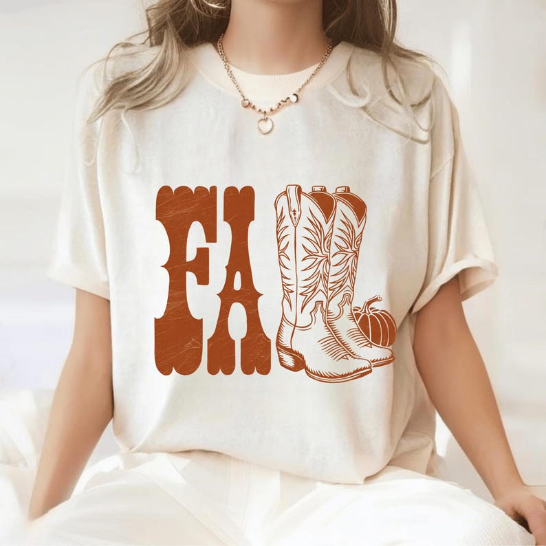 May include: Cream-colored t-shirt featuring the word "FA" in a brown, retro font. The shirt also displays a pair of brown cowboy boots and a pumpkin. The design has a fall or harvest theme.