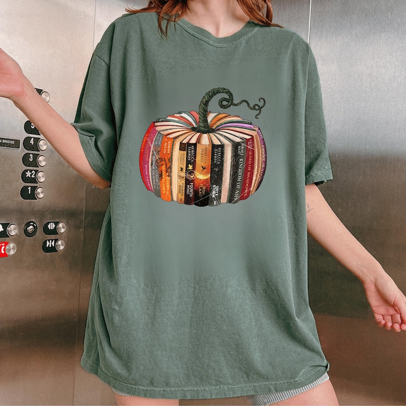 May include: Sage green t-shirt featuring a colorful pumpkin design made of books. The pumpkin has a green stem and is composed of various book spines in shades of red, orange, yellow, and brown. A unique and literary-themed graphic tee.